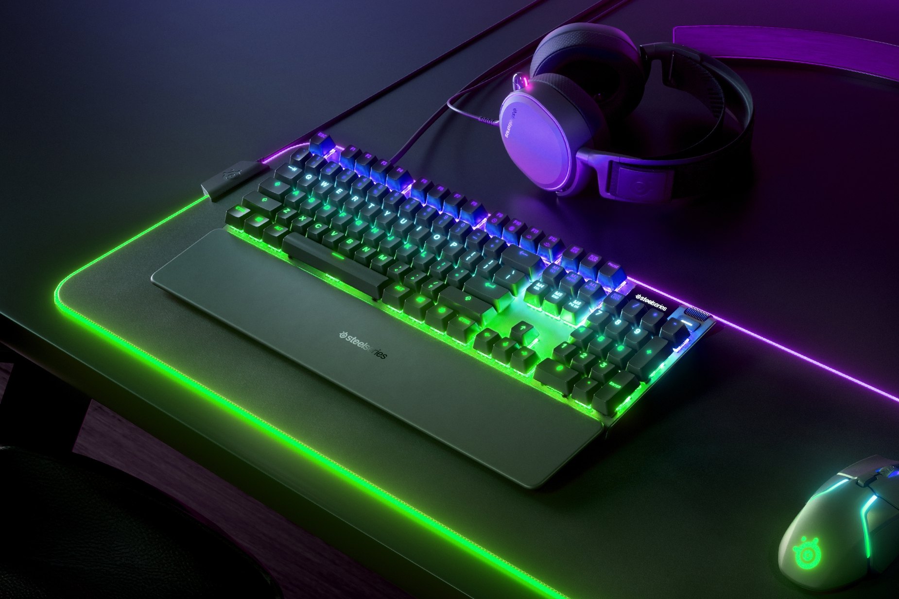 
 UK English - Apex Pro gaming keyboard on a desk with a gaming mouse, both on top of a large mousepad and a SteelSeries gaming headset next to them
 