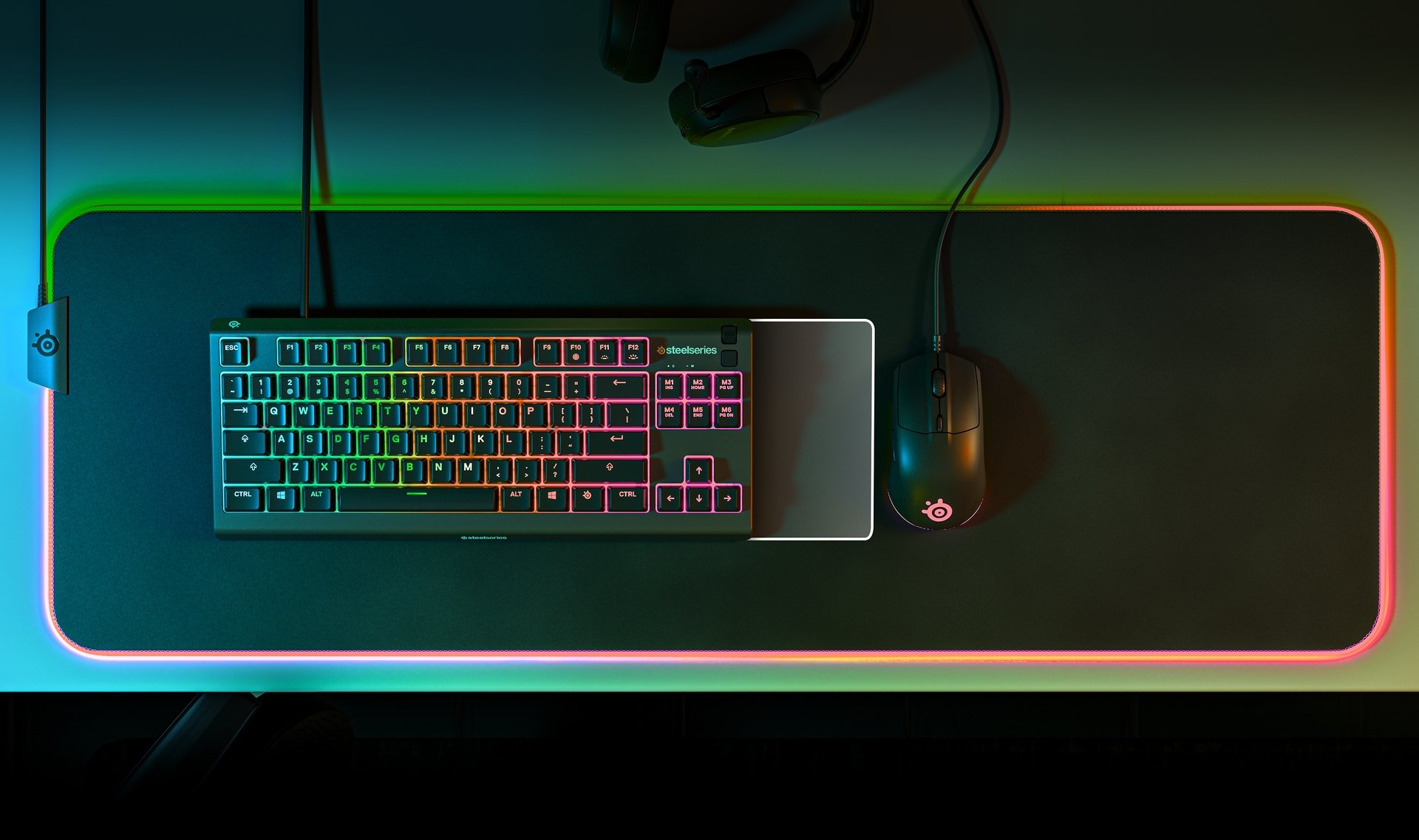 An Apex 3 TKL keyboard sits on an illuminated qck prism cloth mousepade, an Arctis headset, and a SteelSeries mouse. All illuminated in an even glow of RGB.